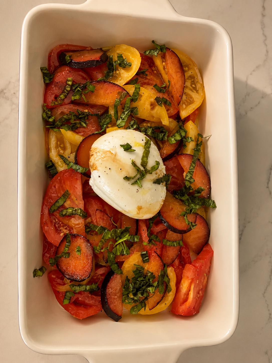 A cream baking dish of heirloom tomatoes and stone fruit under torn burrata