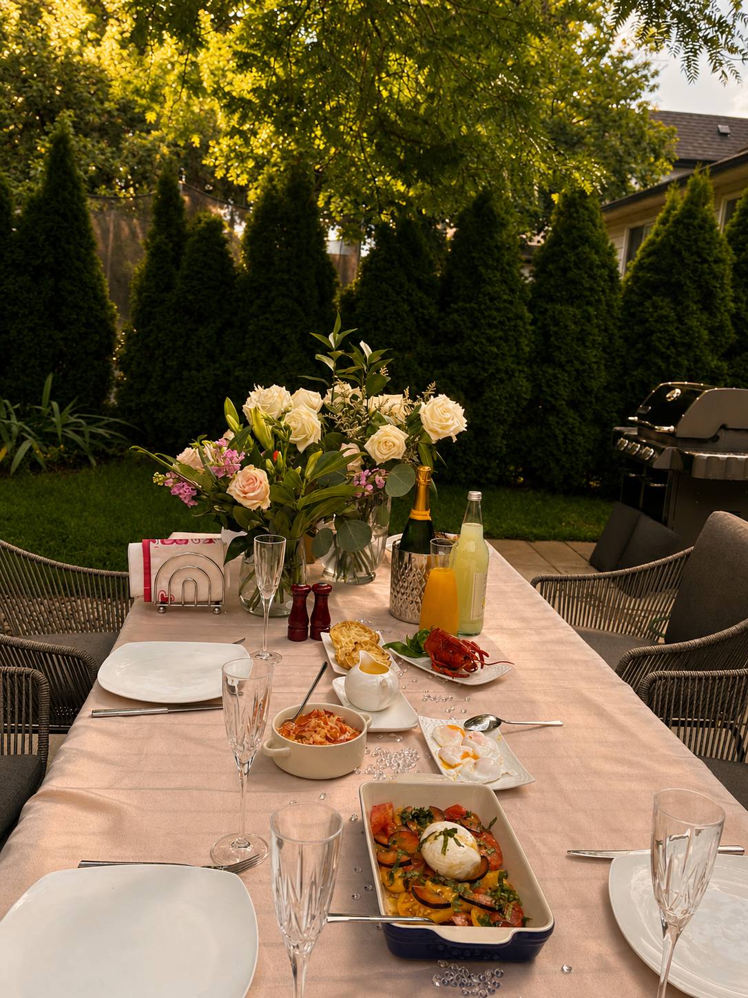 A garden table set with flowers, champagne, and summer dishes under the trees